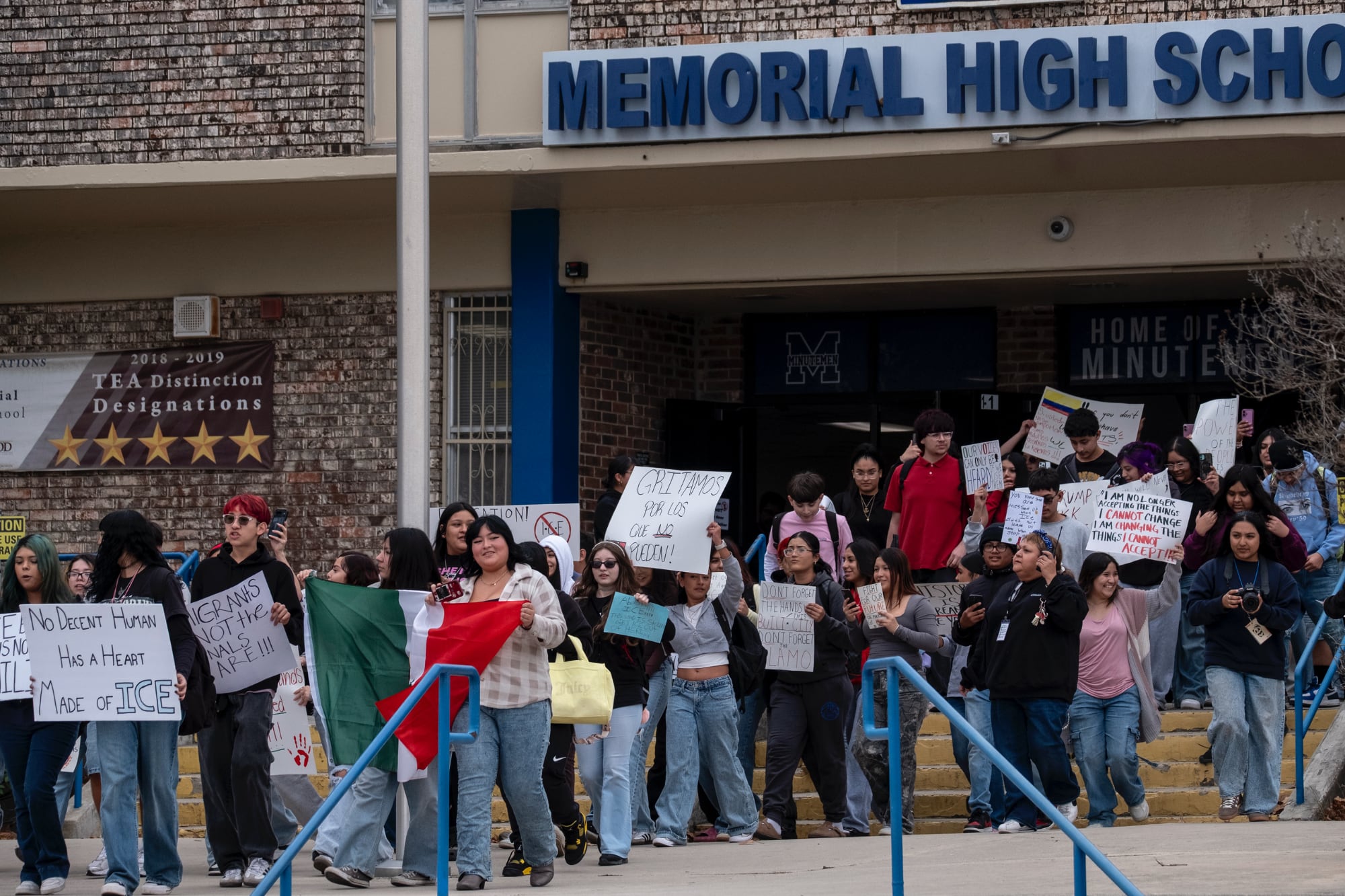 A large group of students at Memorial High School in San Antonio walking out of the building in protest. Several students are holding handmade signs with messages such as "No Decent Human Has a Heart Made of ICE" and "Gritamos por los que no pueden!" while one student prominently displays a Mexican flag.