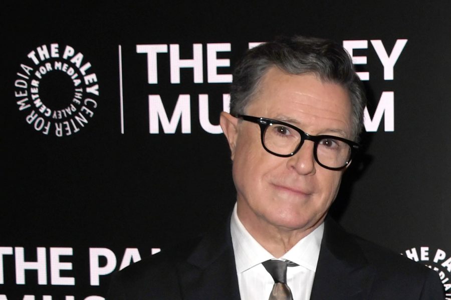 A medium shot of Stephen Colbert wearing a dark suit, white shirt, and patterned tie, posing in front of a black "The Paley Center for Media" backdrop.