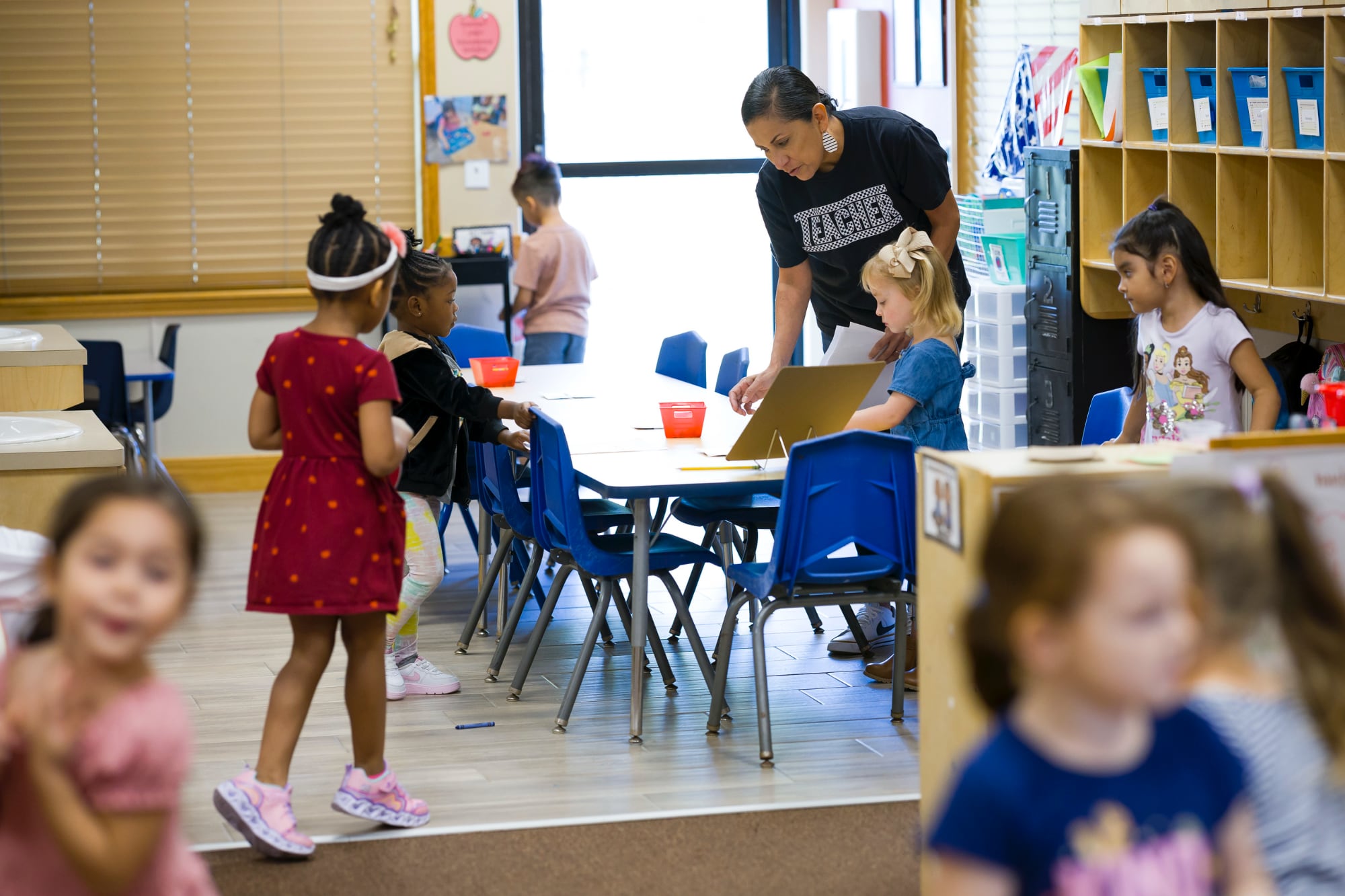 A diverse group of elementary students in a Texas classroom interacting at tables while a teacher in a black shirt leans over to assist a student with instructional materials.