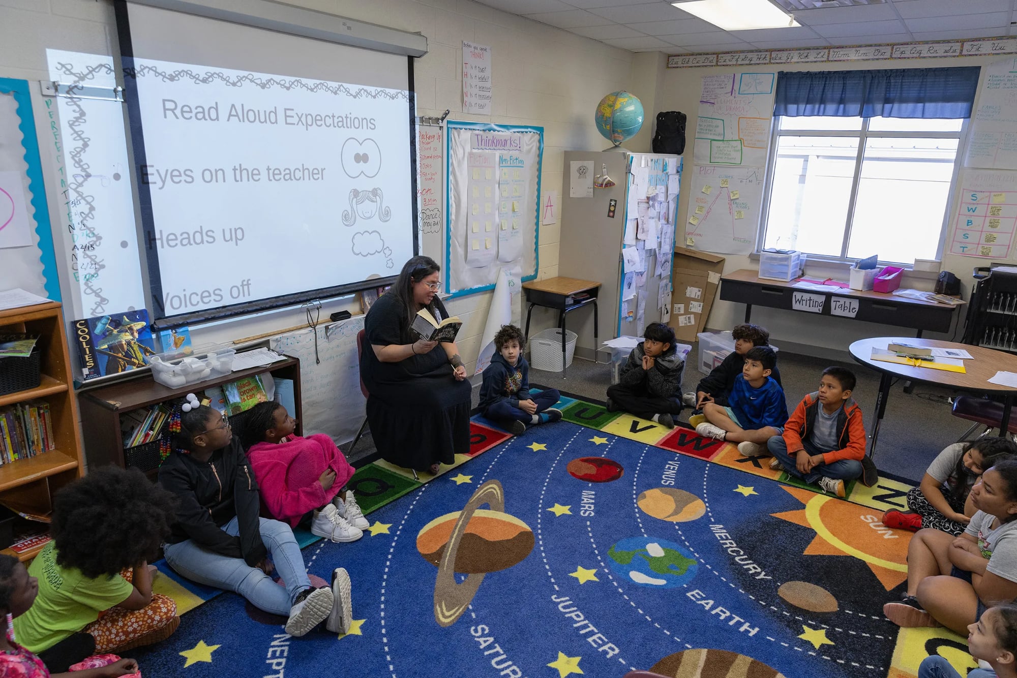 A female teacher sits in a chair reading a book to a diverse group of elementary students seated on a colorful solar system rug in a classroom. A projector screen behind her displays "Read Aloud Expectations," including "Eyes on the teacher," "Heads up," and "Voices off."
