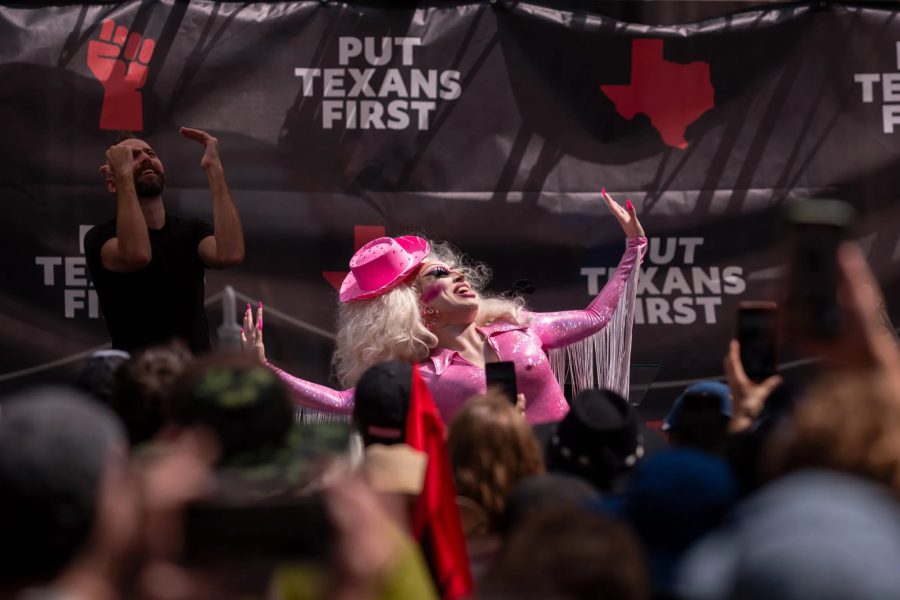 A drag performer in a sparkling pink outfit, blonde wig, and a pink cowboy hat performs on stage at a "Put Texans First" rally. A man to the left of the performer is gesturing with his hands, while a crowd of people in the foreground watches and records the performance on their phones.