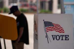 A person in a baseball cap stands at a yellow voting booth in the background, while a white sign in the foreground features an American flag and the word "VOTE" in blue text.
