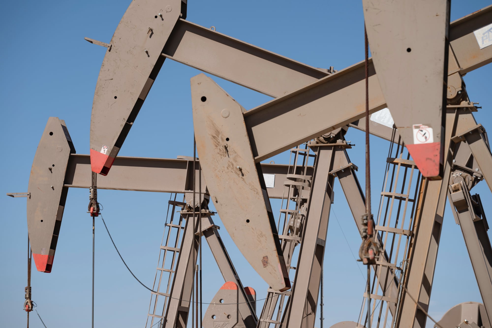 A close-up view of several tan-colored oil pumpjacks operating at a well site in Midland, Texas, against a clear blue sky.
