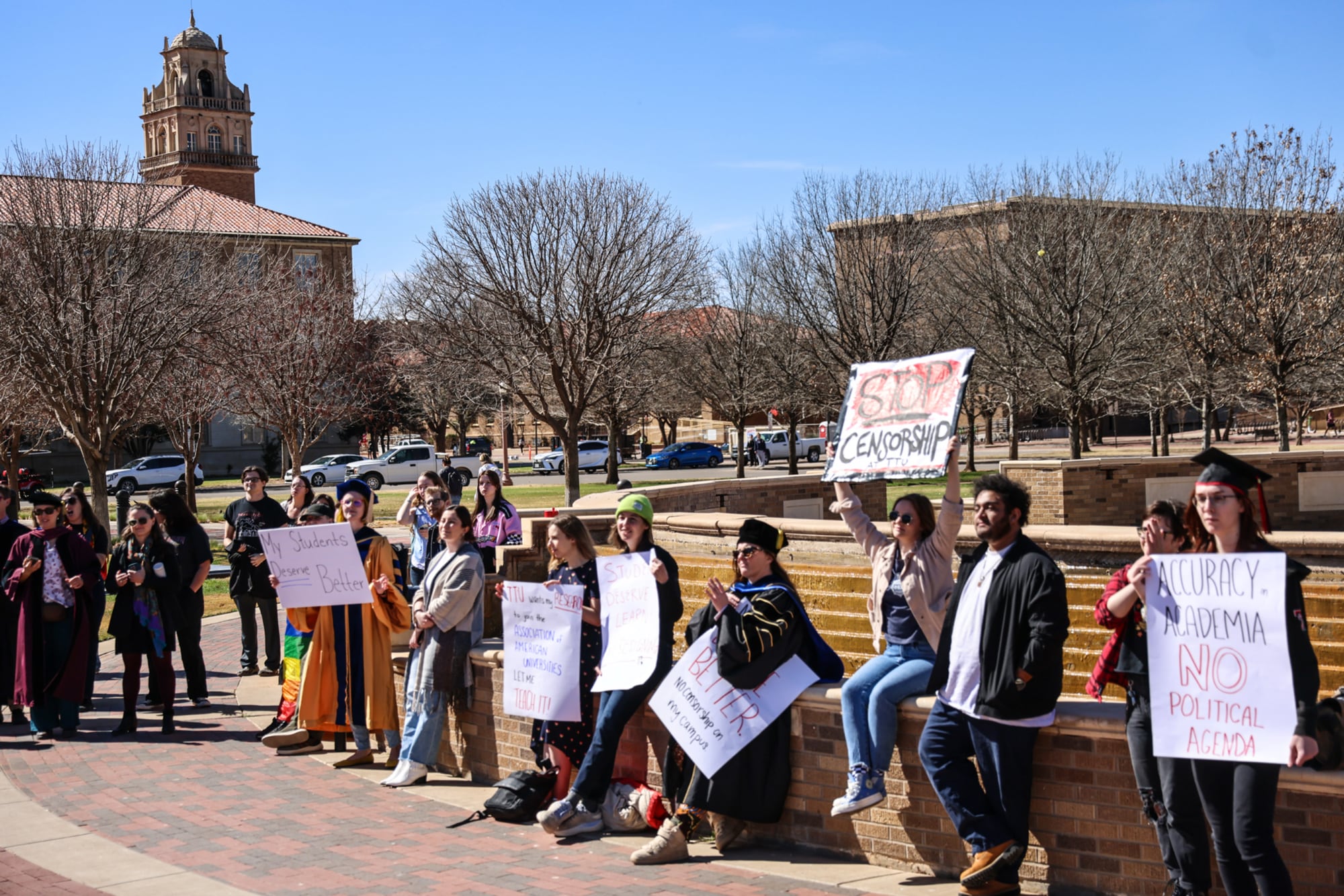 A group of Texas Tech students and faculty members in academic regalia stand and sit near a campus fountain at Memorial Circle, holding handmade protest signs with slogans like "Stop Censorship," "My Students Deserve Better," and "Accuracy in Academia No Political Agenda" during a rally against curriculum changes.