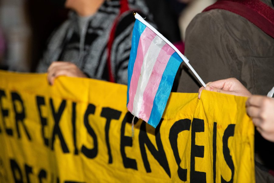 A close-up of a person holding a small blue, pink, and white transgender pride flag over a large yellow protest banner with black hand-painted text.
