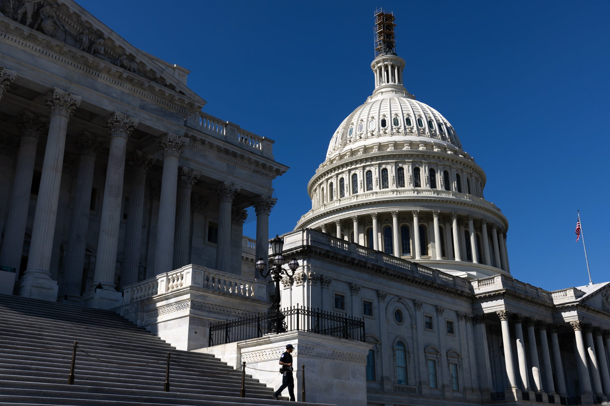 A wide, low-angle shot of the United States Capitol Building under a clear blue sky. The image features the grand marble steps in the foreground, the iconic white dome with scaffolding at the very top, and a single security officer walking near a black lamppost.