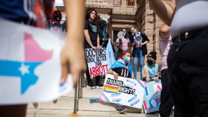 A group of young activists gathered on the steps of the Texas State Capitol during a trans rights rally. One protester holds a sign that reads "Doctors Know Better Than Abbott," while another sits with a hand-painted "Trans Rights" poster decorated with rainbows and butterflies. The scene captures the ongoing tension between student advocates and state leadership over gender identity policies.