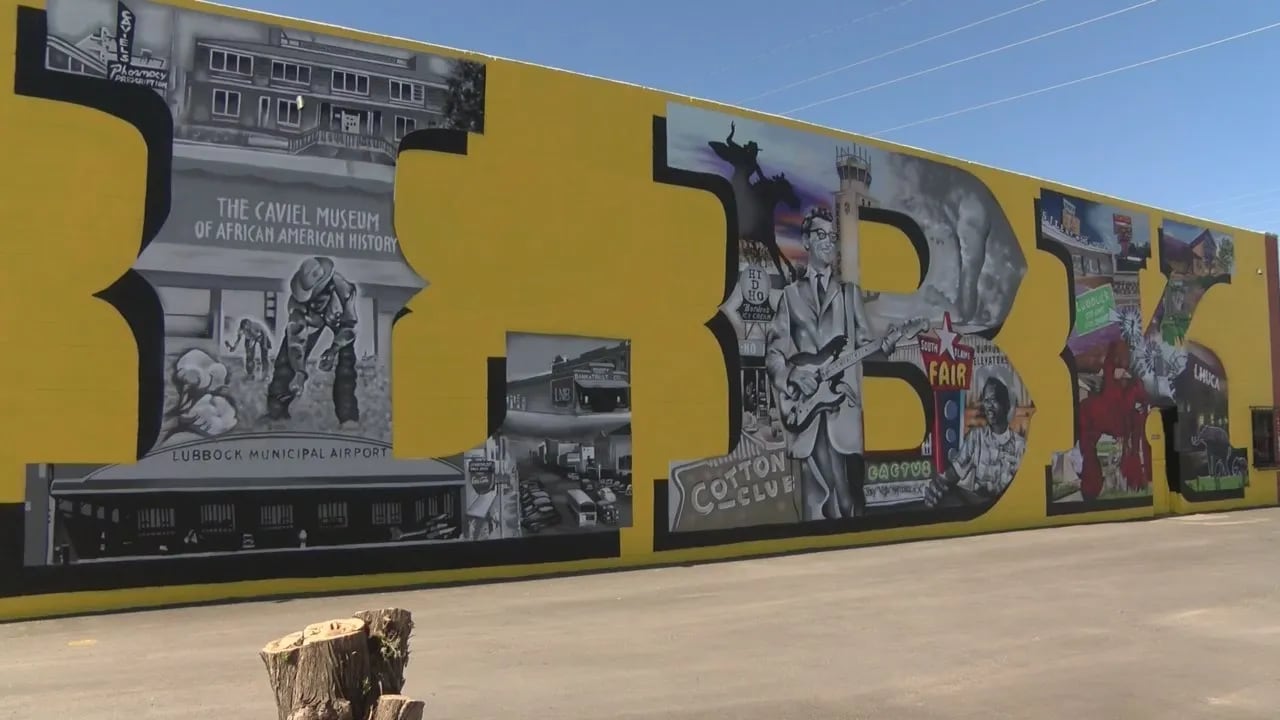 A large outdoor mural on a yellow wall in East Lubbock spells out "LBBK" with historical images like Buddy Holly and the Caviel Museum inside the letters, situated next to a barren asphalt lot.