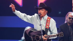 George Strait performing on stage in a cowboy hat and blue plaid shirt, waving to a crowd of 60,000 people who likely skipped their car payments to afford the $3,100 ticket price.