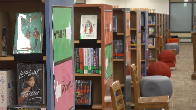 A row of wooden bookshelves in a Lubbock ISD school library featuring colorful "Mystery" and "Romance" signs. Books like "The Love Match" and "Loveboat, Taipei" are prominently displayed on the endcaps, while several grey and red cushioned chairs sit in the background.