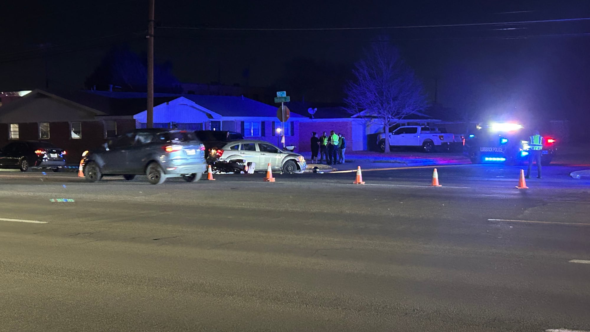 A nighttime view of a crash scene at 53rd and Indiana in Lubbock, Texas, featuring a damaged silver sedan, emergency cones, and police vehicles with flashing lights illuminating a residential neighborhood.
