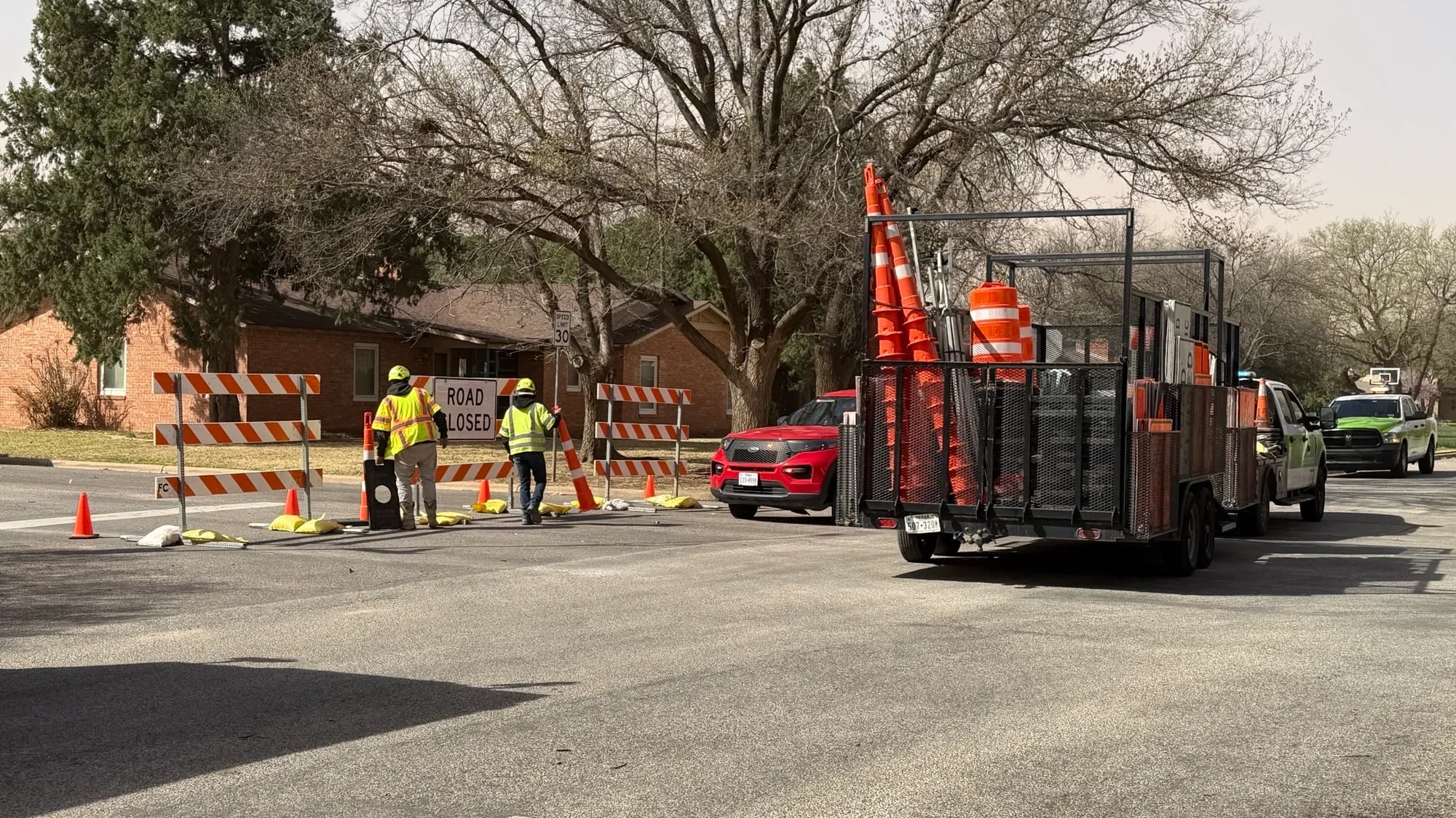Construction workers in neon vests stand around a "Road Closed" sign and orange barricades at the intersection of 18th and Toledo in Lubbock, Texas. A trailer overflowing with more cones sits in the foreground, while the sky in the background is a hazy, dusty shade of "West Texas Brown" thanks to 50 mph wind gusts that are currently mocking our city’s fragile infrastructure.