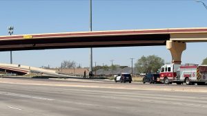An emergency scene under a concrete Lubbock overpass (labeled with a '16 FT 5 IN' sign). A marked police SUV with flashing lights, a fire engine, and responders stand on the shoulder. On the on-ramp lane above them, a lone motorcycle lies on its side, the site of a single motorcycle crash, on a clear day. A cell tower is visible in the distance.
