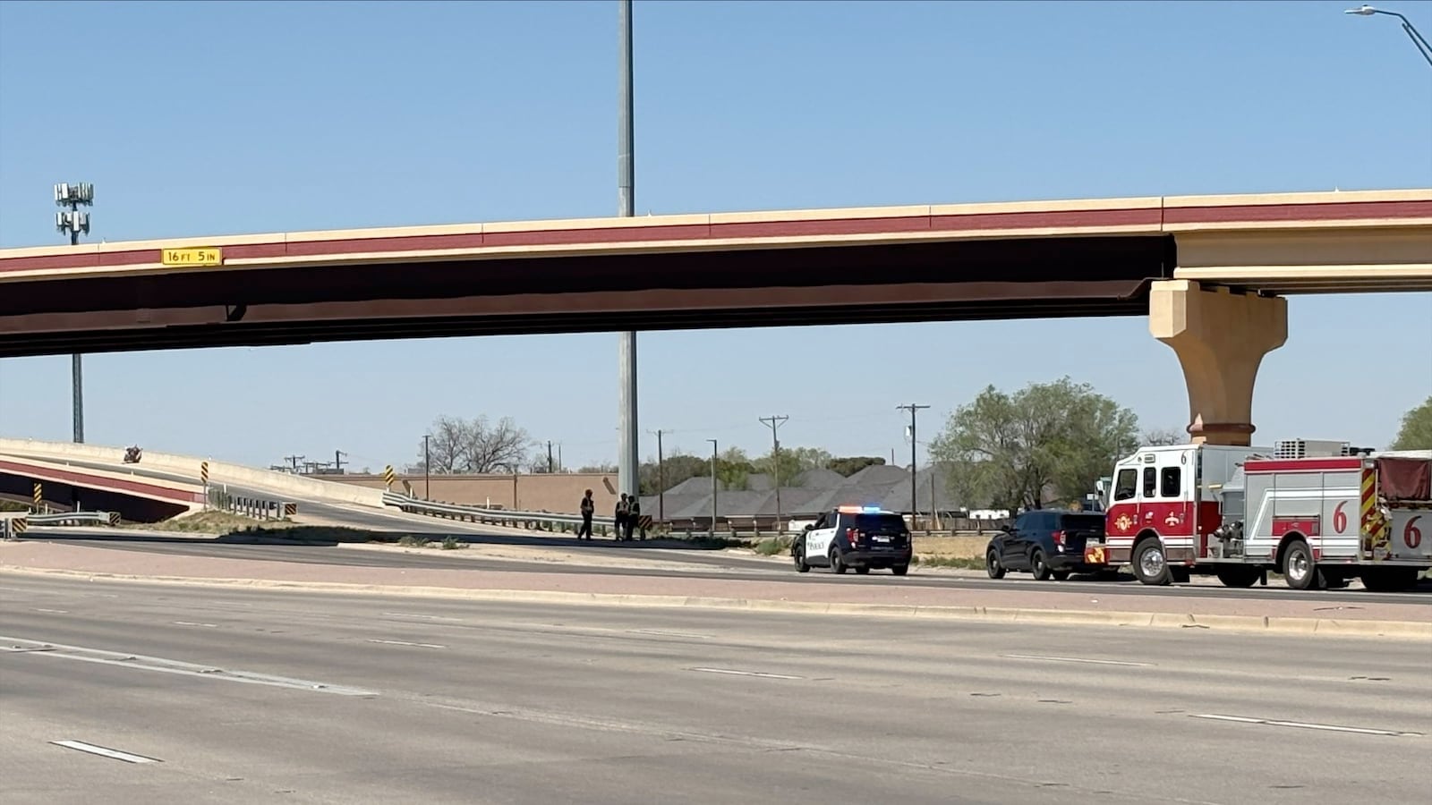 An emergency scene under a concrete Lubbock overpass (labeled with a '16 FT 5 IN' sign). A marked police SUV with flashing lights, a fire engine, and responders stand on the shoulder. On the on-ramp lane above them, a lone motorcycle lies on its side, the site of a single motorcycle crash, on a clear day. A cell tower is visible in the distance.