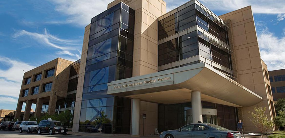 The exterior of the Texas Tech Physicians Medical Pavilion in Lubbock, Texas, featuring a sleek glass tower with a blue DNA double helix design that serves as a beautiful reminder of exactly whose genetic code will be used to collateralize your new medical debt.