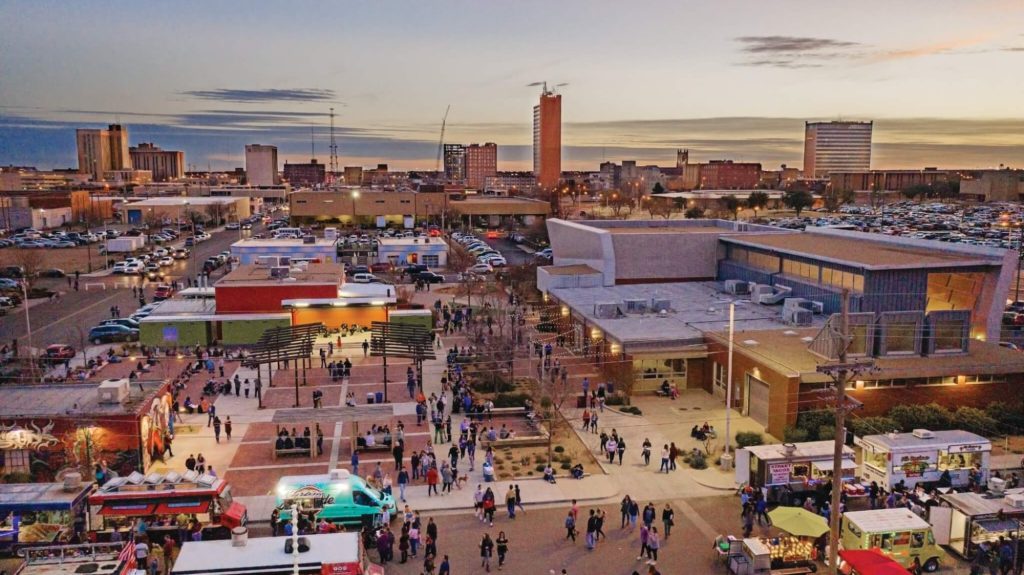An aerial drone view of the First Friday Art Trail at the LHUCA campus, capturing the rare three-hour window where downtown Lubbock actually contains signs of life. Food trucks and optimistic locals gather in the plaza while the West Texas sunset tries its hardest to make the 30-year "revitalization" project look like it’s finished.