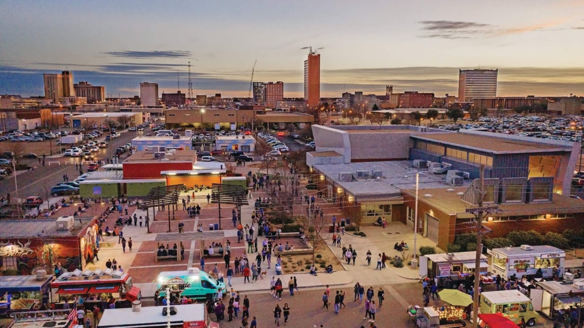 An aerial drone view of the First Friday Art Trail at the LHUCA campus, capturing the rare three-hour window where downtown Lubbock actually contains signs of life. Food trucks and optimistic locals gather in the plaza while the West Texas sunset tries its hardest to make the 30-year "revitalization" project look like it’s finished.