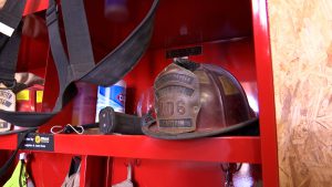 A weathered brown leather firefighter captain's helmet with a "West Carlisle Fire Dept 106" shield sits on a bright red metal shelf next to a container of Clorox wipes and firefighting gear.