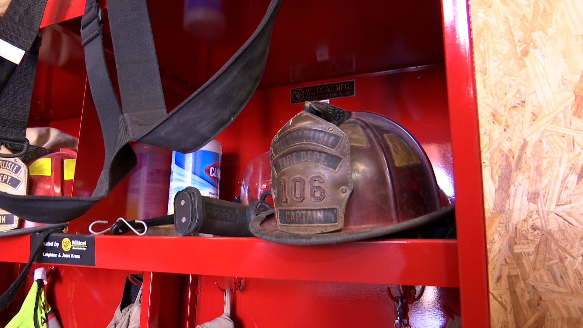 A weathered brown leather firefighter captain's helmet with a "West Carlisle Fire Dept 106" shield sits on a bright red metal shelf next to a container of Clorox wipes and firefighting gear.