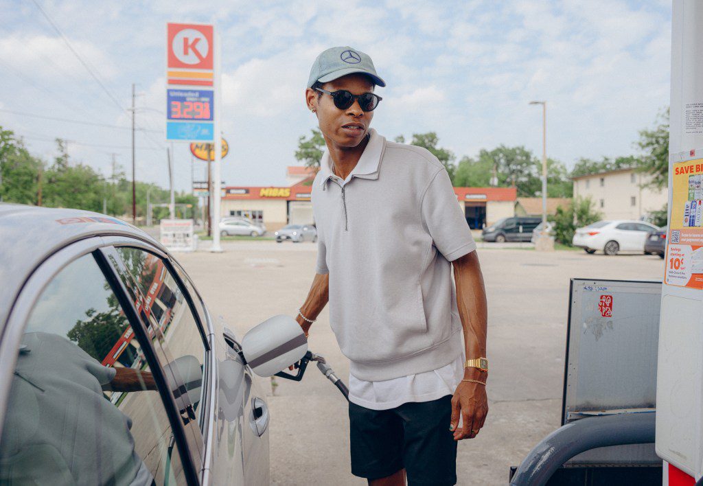 A young man stares blankly into the middle distance while fueling his car, contemplating if walking ten miles in the Lubbock wind is actually a viable lifestyle choice.