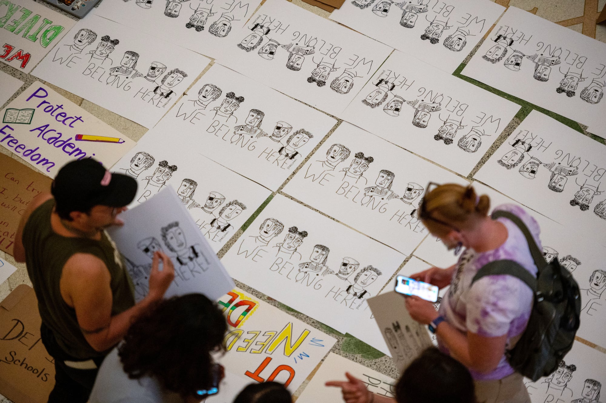 An overhead shot of the Texas Capitol rotunda floor covered in student-made posters that say "WE BELONG HERE" in an optimistic attempt to explain basic human dignity to a state government that is currently building a "woke" snitch-line without a manual.