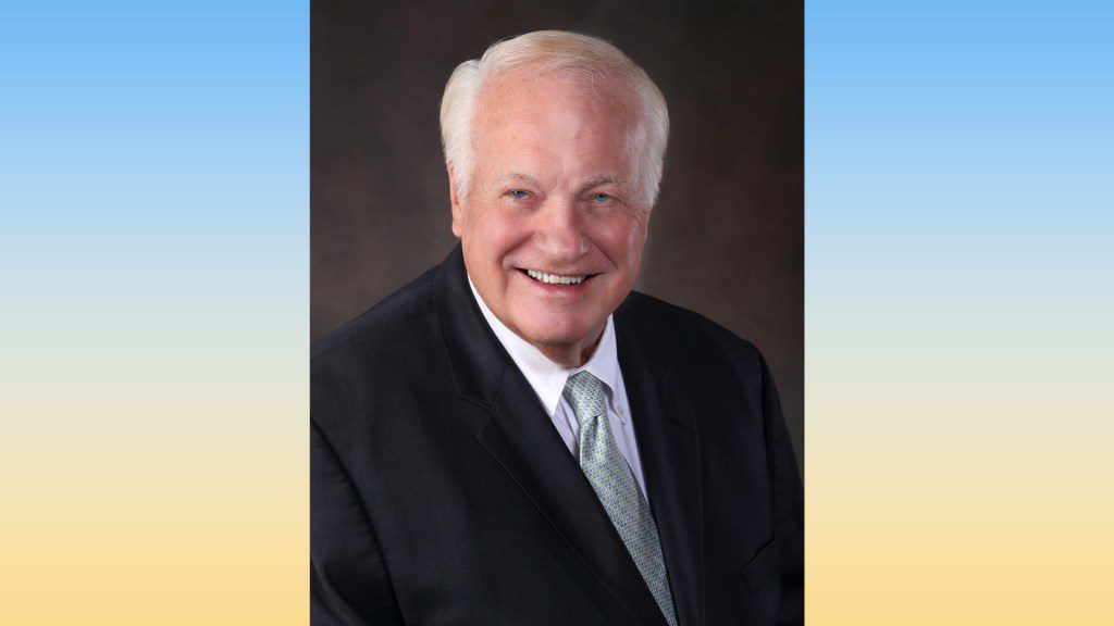 A headshot of Gary Boren, an older man with white hair wearing a suit and tie, smiling against a blue and yellow gradient background.