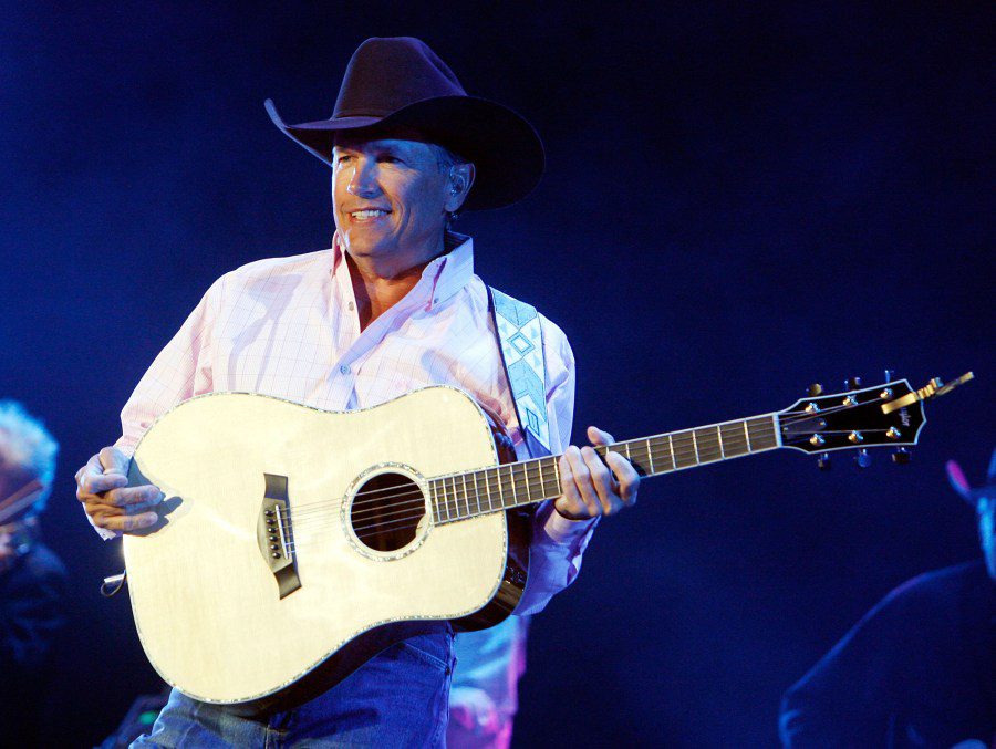 George Strait smiling and playing an acoustic guitar while wearing a purple cowboy hat and a pink checkered shirt.