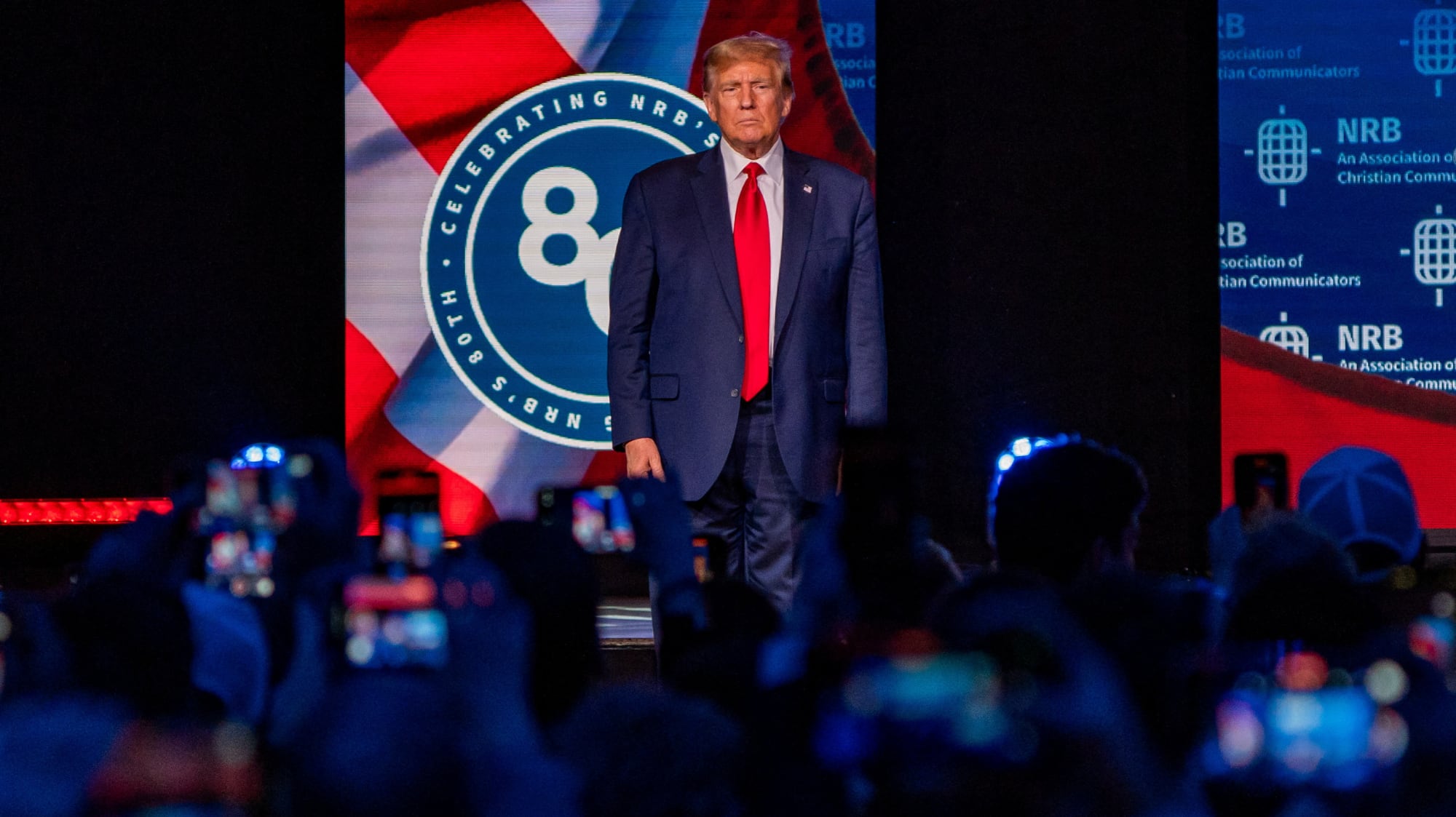 Donald Trump stands on a stage at the NRB convention, framed by Christian media logos and a sea of glowing smartphones, looking exactly like the guy your Lubbock aunt wants to hear a sermon from instead of actually reading the Gospels.