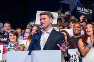 State Representative James Talarico gestures while speaking at a podium, surrounded by supporters holding signs that read "Talarico for Texas."