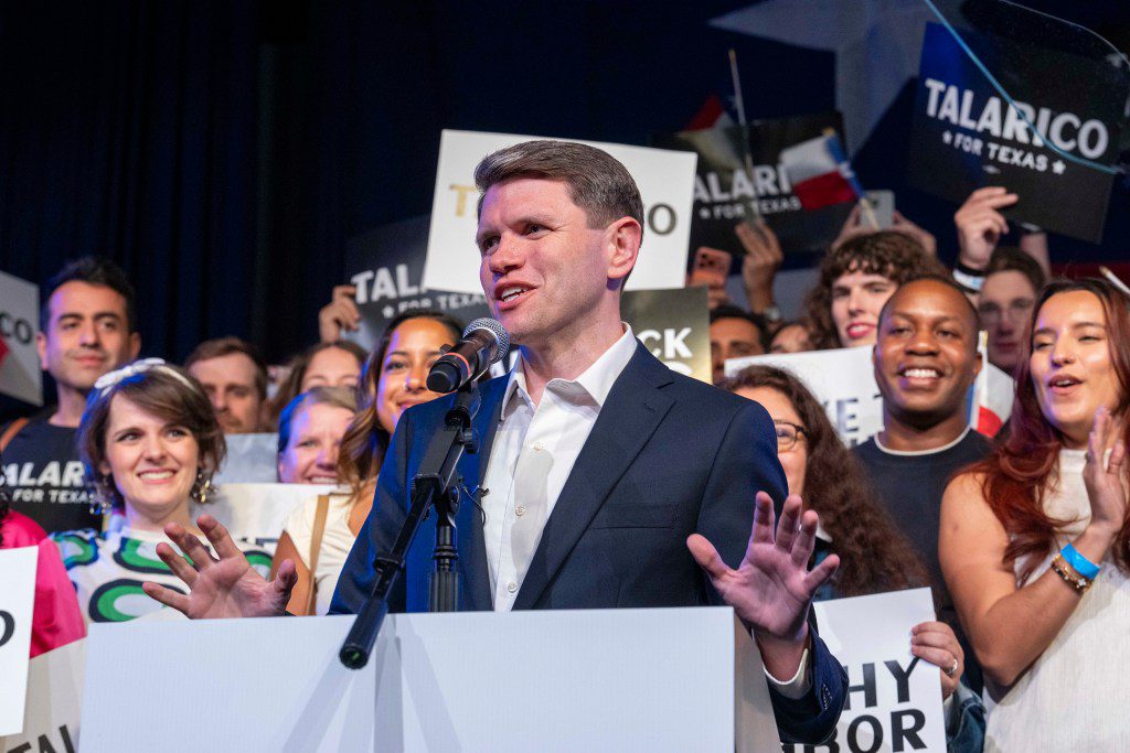State Representative James Talarico gestures while speaking at a podium, surrounded by supporters holding signs that read "Talarico for Texas."
