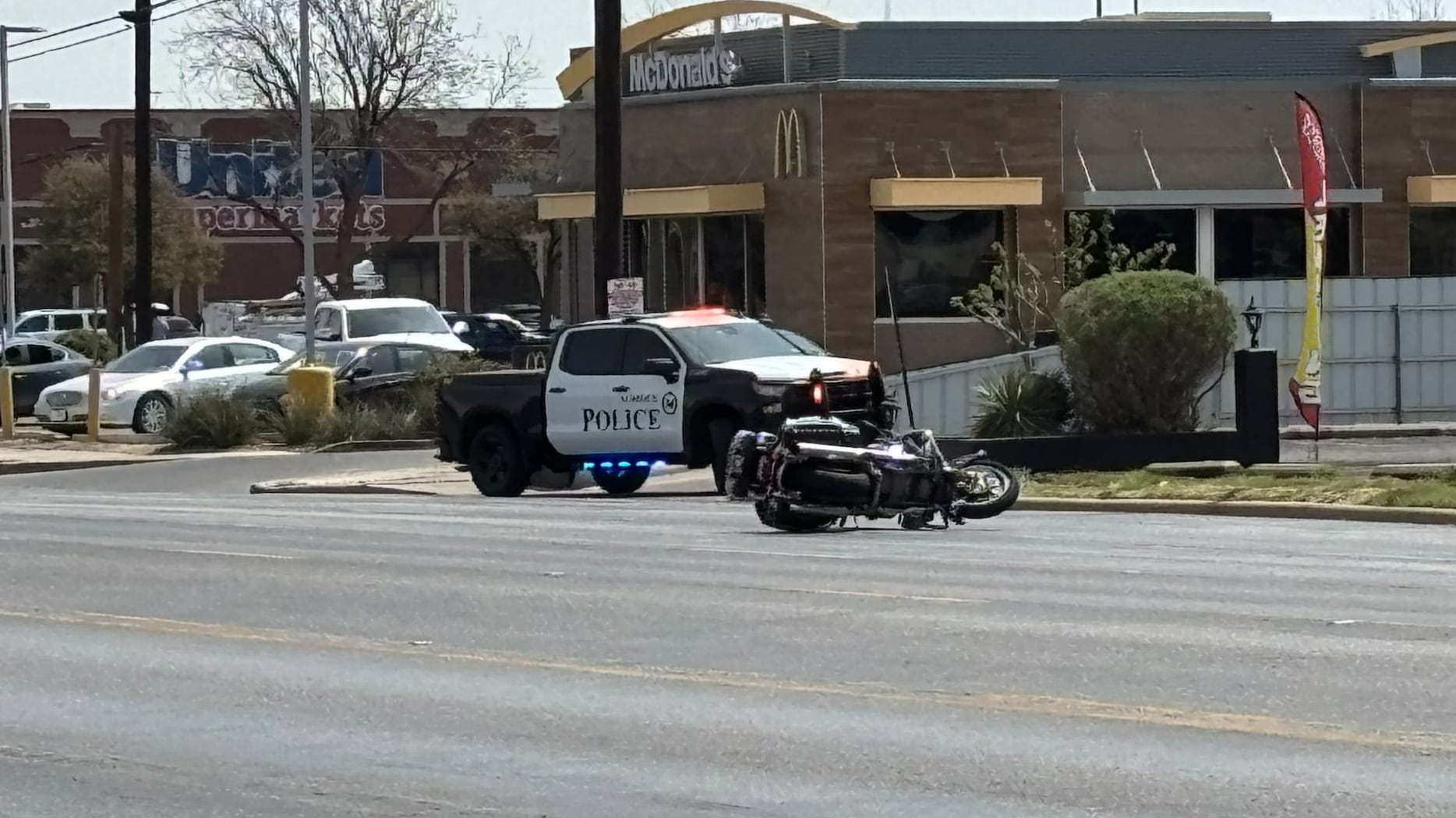 A crashed Lubbock Police motorcycle lying horizontally on the asphalt of 4th Street, perfectly positioned in front of a McDonald's to highlight the exact moment bravery met a "Do Not Pursue" order.