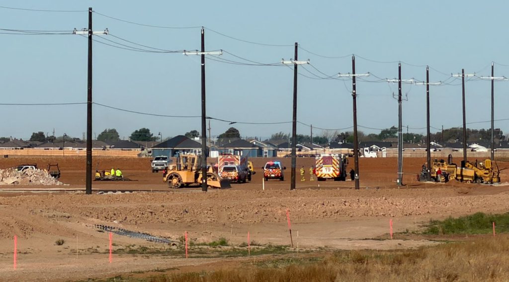 A flat, dusty construction site at 114th and Upland featuring a front-end loader, emergency vehicles with flashing lights, and a row of utility poles, all set against a backdrop of Lubbock’s finest generic suburban sprawl and endless brown dirt.