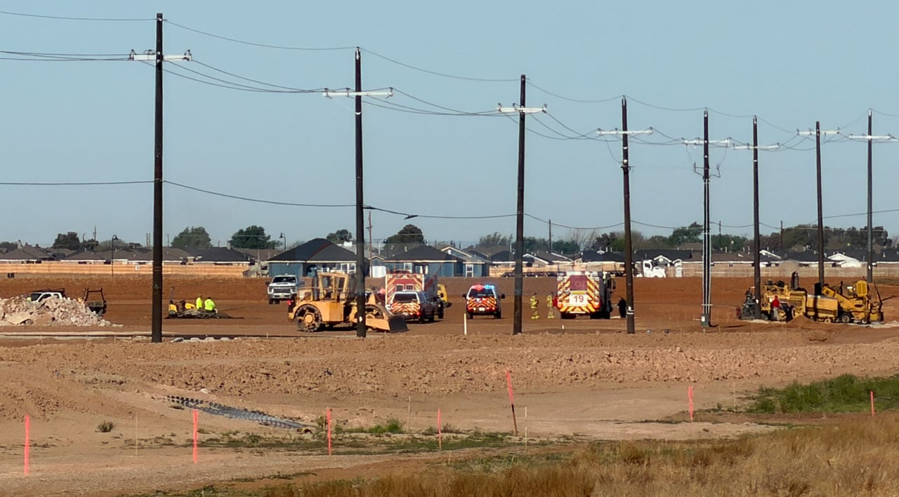 A flat, dusty construction site at 114th and Upland featuring a front-end loader, emergency vehicles with flashing lights, and a row of utility poles, all set against a backdrop of Lubbock’s finest generic suburban sprawl and endless brown dirt.