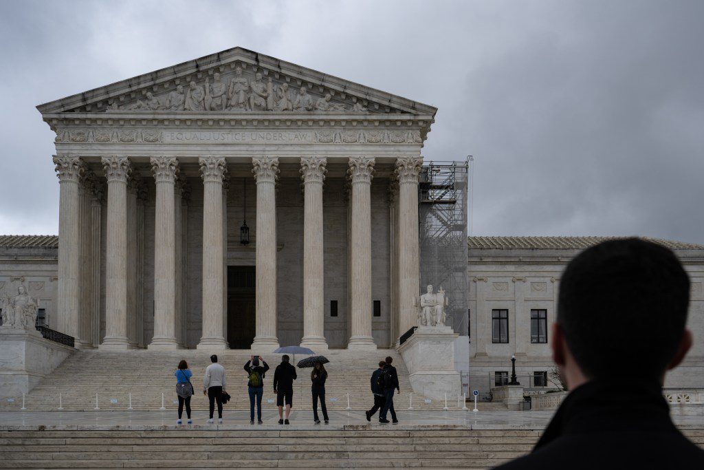 The U.S. Supreme Court building looms under a gloomy, overcast sky while tourists take photos on the marble steps.
