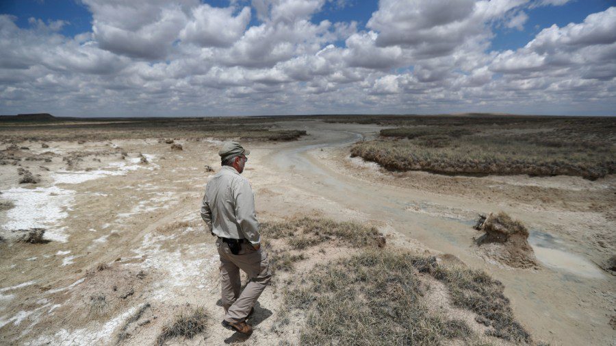 A man in a tan shirt and tactical pants stands in a desolate, dry West Texas landscape looking over a muddy creek bed under a cloudy sky.