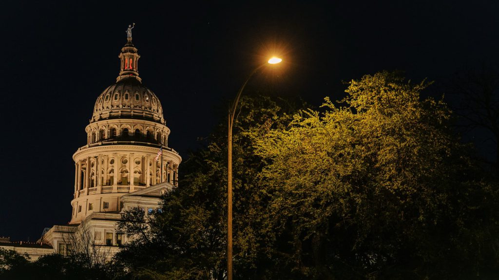 The dome of the Texas State Capitol building illuminated at night in Austin, seen from behind a large, leafy tree glowing under a streetlamp.