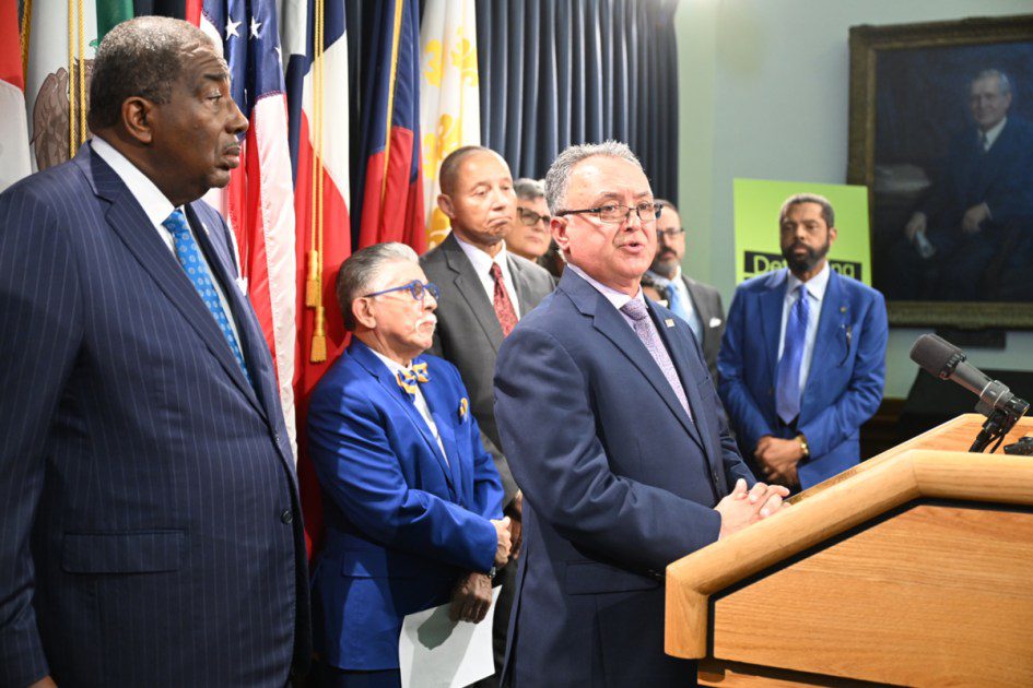A group of men in business suits, including state officials and legal representatives, gather at a wooden podium for a press conference regarding the Historically Underutilized Business program lawsuit.