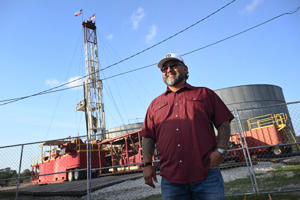 A man in a maroon shirt and baseball cap stands in front of a drilling rig under a clear Texas sky, symbolizing the state's desperate search for resources.