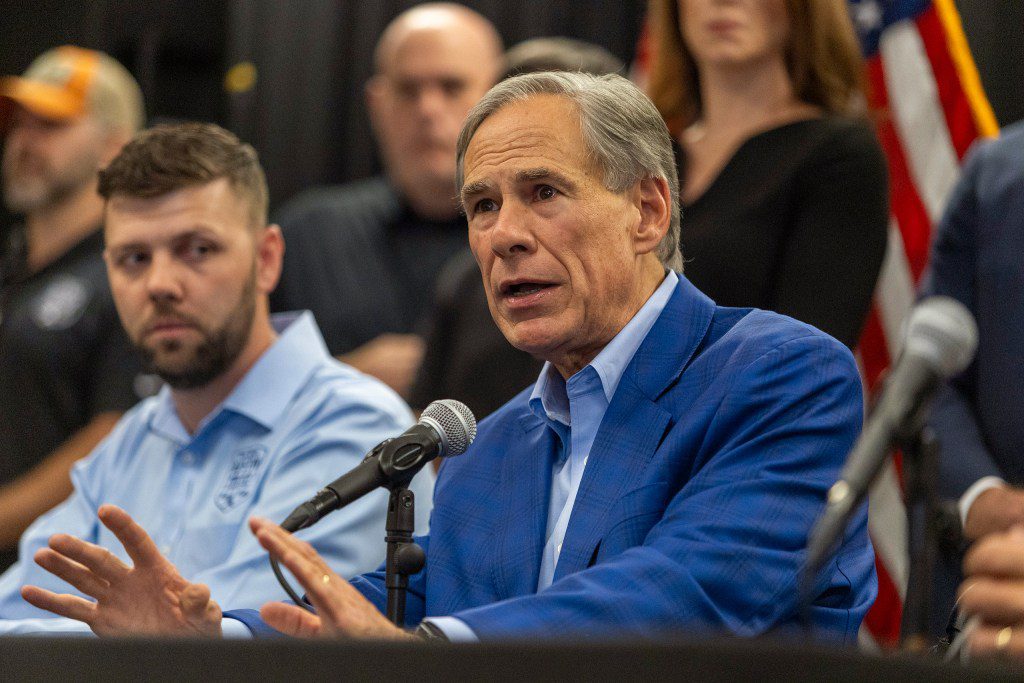 Texas Governor Greg Abbott speaking into a microphone at a press conference table alongside other officials.