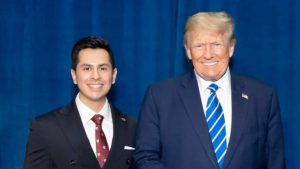 Congressional candidate Abraham Enriquez in a dark suit standing next to a smiling Donald Trump, who is giving a thumbs-up in front of a blue backdrop.