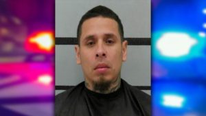 Police mugshot of Arthur Soto with a goatee and neck tattoos, set against a graphic background of blurred red and blue police emergency lights.