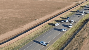 An aerial view of multiple police cruisers and emergency vehicles blocking a two-lane rural Texas highway next to a barren, flat dirt field.