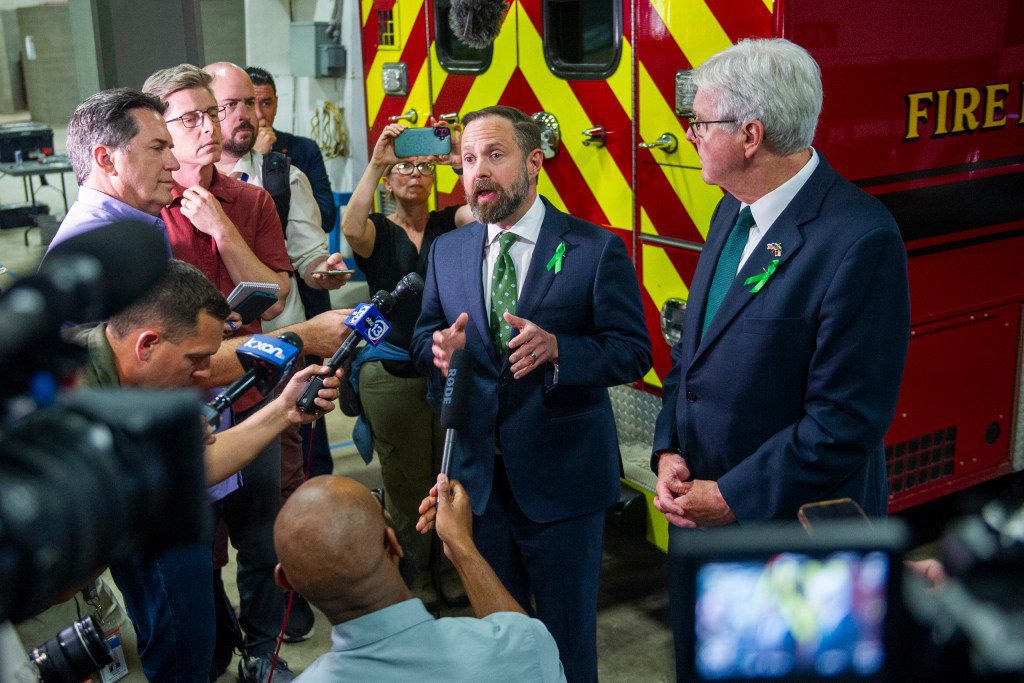 Texas House Speaker Dustin Burrows and Lieutenant Governor Dan Patrick speaking to reporters at a press conference in front of a red fire truck, wearing green awareness ribbons.