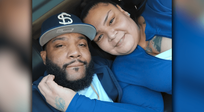 A selfie of Jessica Baker smiling next to her husband, Khadafi 'Michael' Baker, who is wearing a blue baseball cap with an interlocking 'S' logo.