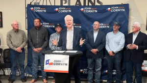 Senator John Cornyn speaking at a John Cornyn for Senate campaign podium in Lubbock, flanked by supporters in button-down shirts against a blue backdrop.