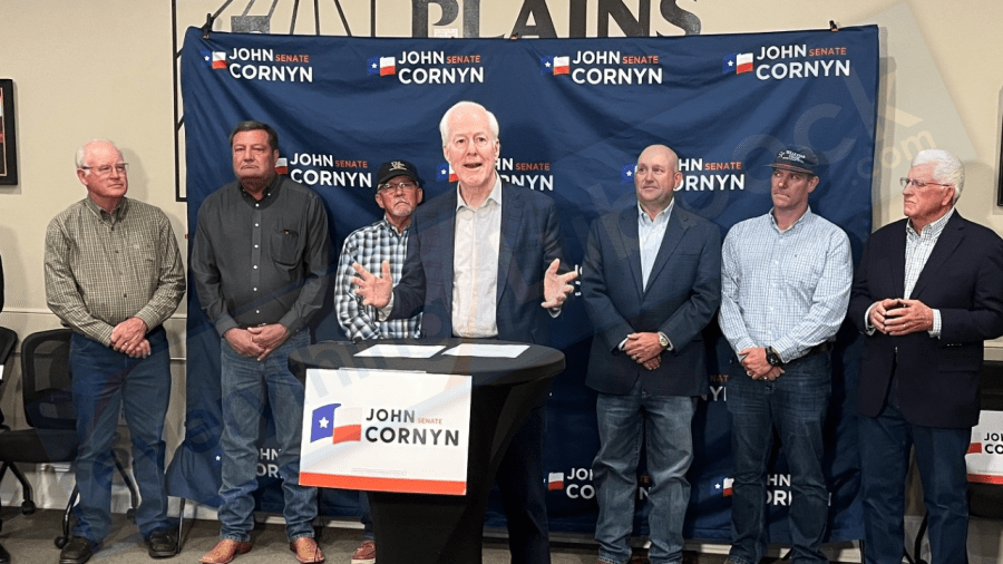 Senator John Cornyn speaking at a John Cornyn for Senate campaign podium in Lubbock, flanked by supporters in button-down shirts against a blue backdrop.