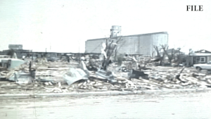 A grainy black-and-white archival photo showing the widespread destruction of the 1970 Lubbock tornado, with flattened homes in the foreground and a large grain elevator standing in the distance.