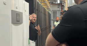A man in a black shirt gestures toward white Schneider Electric server enclosures while giving a presentation to a group of onlookers inside a data center corridor.