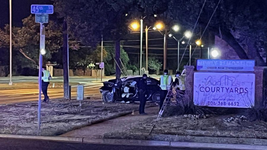 A heavily damaged car sits wrecked on the grass next to the "Courtyards" apartment complex sign at 45th and University at night, with Lubbock police officers and flashing lights surrounding the scene.