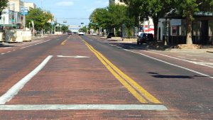 A view down the historic red-brick paved Broadway street in downtown Lubbock, showing yellow lines and a left-turn arrow painted on the bumpy surface.